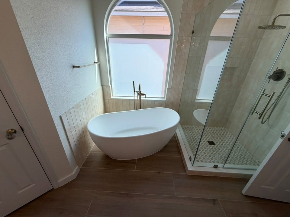 Freestanding soaking tub beneath an arched window, beside a glass walk-in shower, on wood-look floor