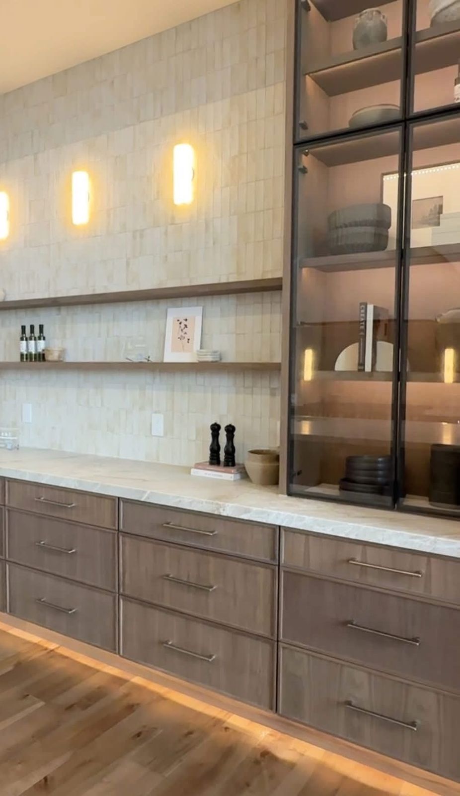 Kitchen remodel with wood-tone flat-panel drawers, a stone countertop, stacked tile wall with sconces and floating shelves, and lit glass-front display cabinets