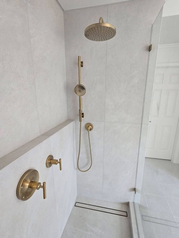 White tile shower with brushed brass rain head, handheld shower and valve, and a linear drain
