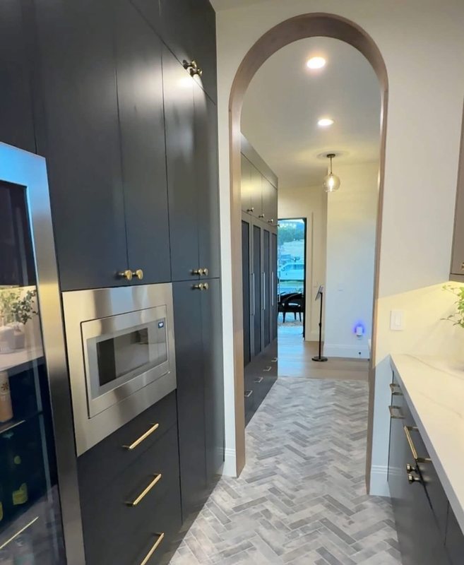 Dark floor-to-ceiling cabinetry with built-in wall ovens beside a herringbone tile hallway and arched opening
