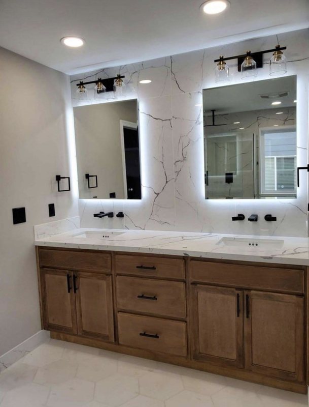 Wood double vanity with a white countertop, two backlit LED mirrors, black faucets and a marble slab accent wall